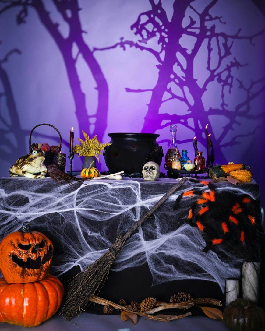 A Halloween-themed table with a cauldron, skull, candles, potions, pumpkins, a broom, and fake spider webs, set against a purple background with eerie tree branch shadows.