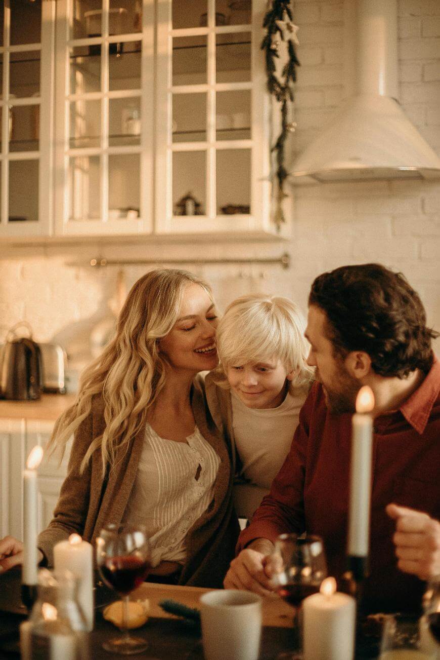 A woman, a man, and a young boy sit together at a candlelit table in a cozy kitchen, smiling and enjoying each other’s company. The atmosphere is warm and intimate, with soft lighting and wine glasses on the table.