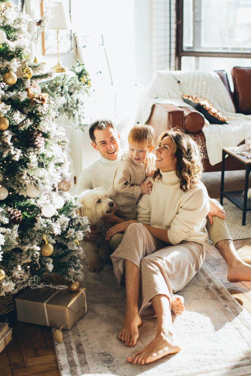 A smiling family of three and their dog sit on the floor by a decorated Christmas tree, surrounded by sunlight and wrapped gifts in a cozy living room.