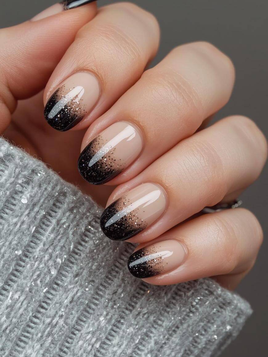 A close-up of a hand with manicured nails featuring a nude base and black glitter ombre tips, against a silver-gray, ribbed, and sparkly sweater. The nails are almond-shaped and glossy.