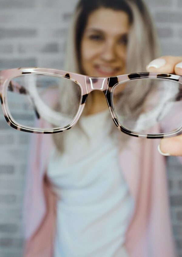 A person holds a pair of eyeglasses toward the camera, with the glasses in sharp focus and the persons face and body blurred in the background against a light brick wall.