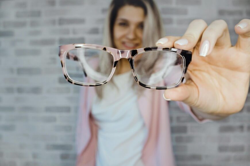 A person holding a pair of eyeglasses close to the camera, with their face out of focus in the background against a light brick wall.