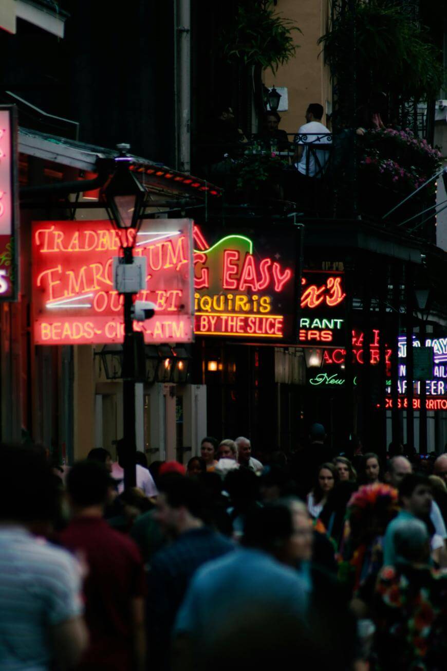 A crowded street scene at dusk with colorful neon signs for bars and restaurants glowing above the heads of many people walking below. Some signs read EMERIUM, EASY, and BEADS.