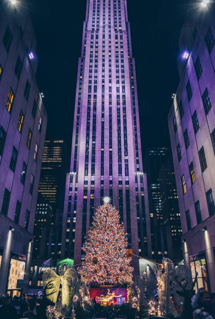 A tall Christmas tree covered in colorful lights stands in front of a high-rise building at night, surrounded by festive decorations and crowds, in a city plaza illuminated with purple lighting.