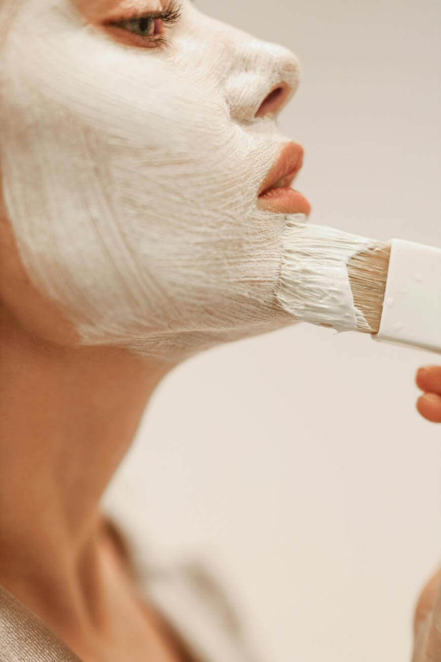Close-up of a person applying a white facial mask with a brush to their face, focusing on the lower cheek and chin area. The background is softly blurred.
