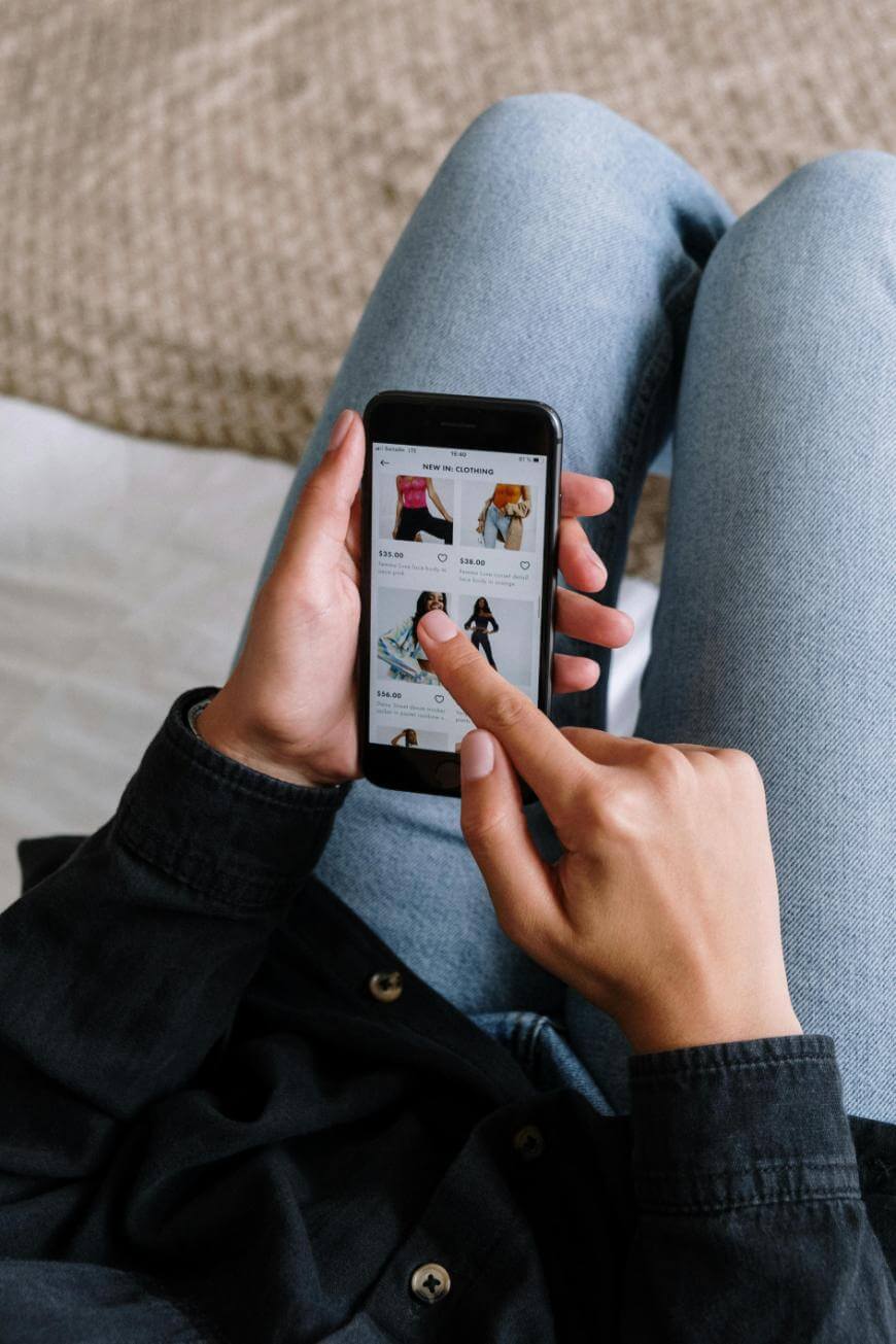 A person sitting with legs crossed, holding a smartphone and browsing an online clothing store, scrolling through various apparel options on the screen.