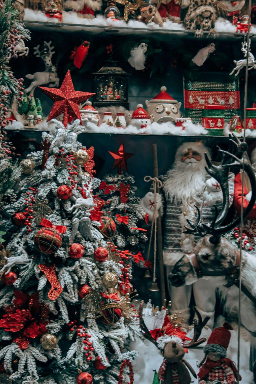A festive Christmas display features a decorated tree with red and gold ornaments, Santa Claus figures, reindeer, and various holiday decorations, all surrounded by artificial snow.