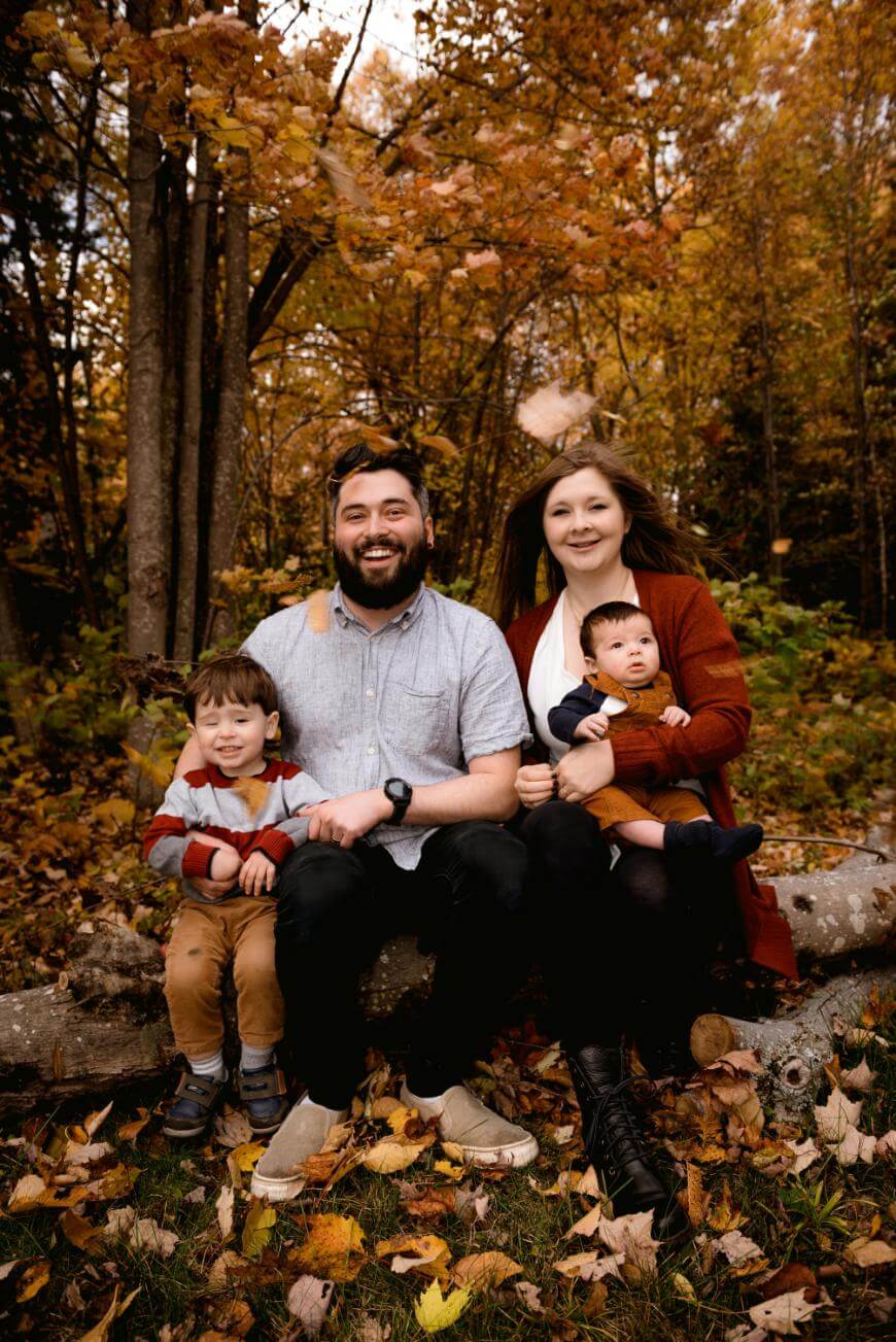 A family of four sits on a log outdoors in autumn, surrounded by colorful fall leaves. The parents smile, with a young boy sitting beside them and a baby in the mothers lap. Trees with orange foliage are in the background.