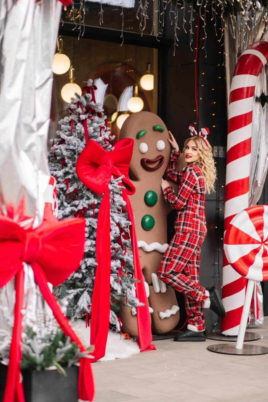 A woman in red plaid pajamas stands next to a large gingerbread man decoration by a snowy Christmas tree with big red bows, surrounded by festive holiday decorations, including oversized candy canes and peppermint candies.