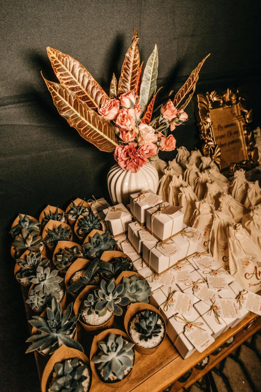 A wooden table holds small succulent plants in pots, white gift boxes tied with twine, cream-colored drawstring bags, and a white vase with pink roses and gold-tinged foliage against a dark background.