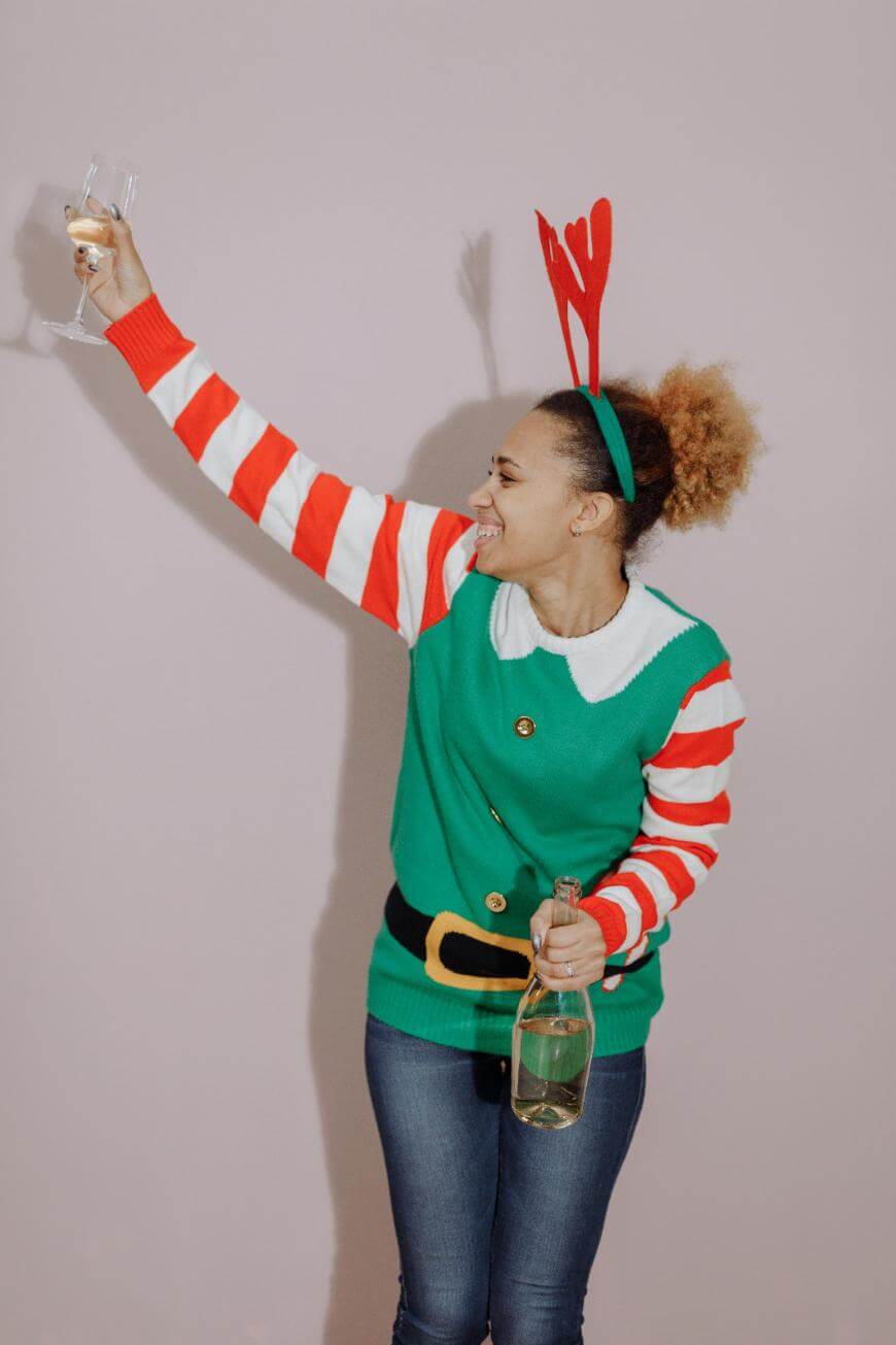 A smiling person wearing a festive elf sweater and antler headband raises a champagne glass in one hand and holds a bottle of champagne in the other, celebrating against a plain light-colored background.