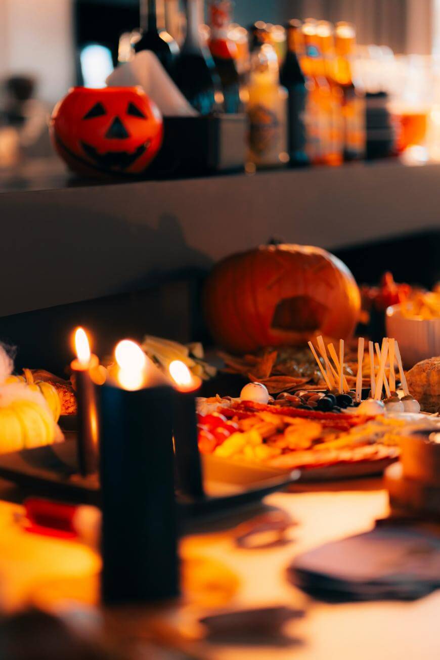 A festive table features Halloween decorations, including carved pumpkins and lit black candles, with various snacks and drinks arranged for a party.