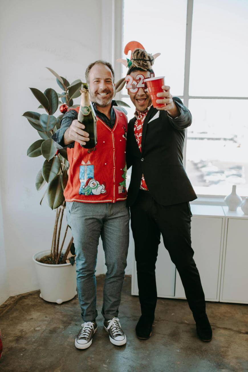 Two men smile and toast with drinks at a festive party. One wears a red holiday sweater and holds champagne; the other wears a suit, red cup, and a festive headband with “23.” They stand by a plant in a bright room.
