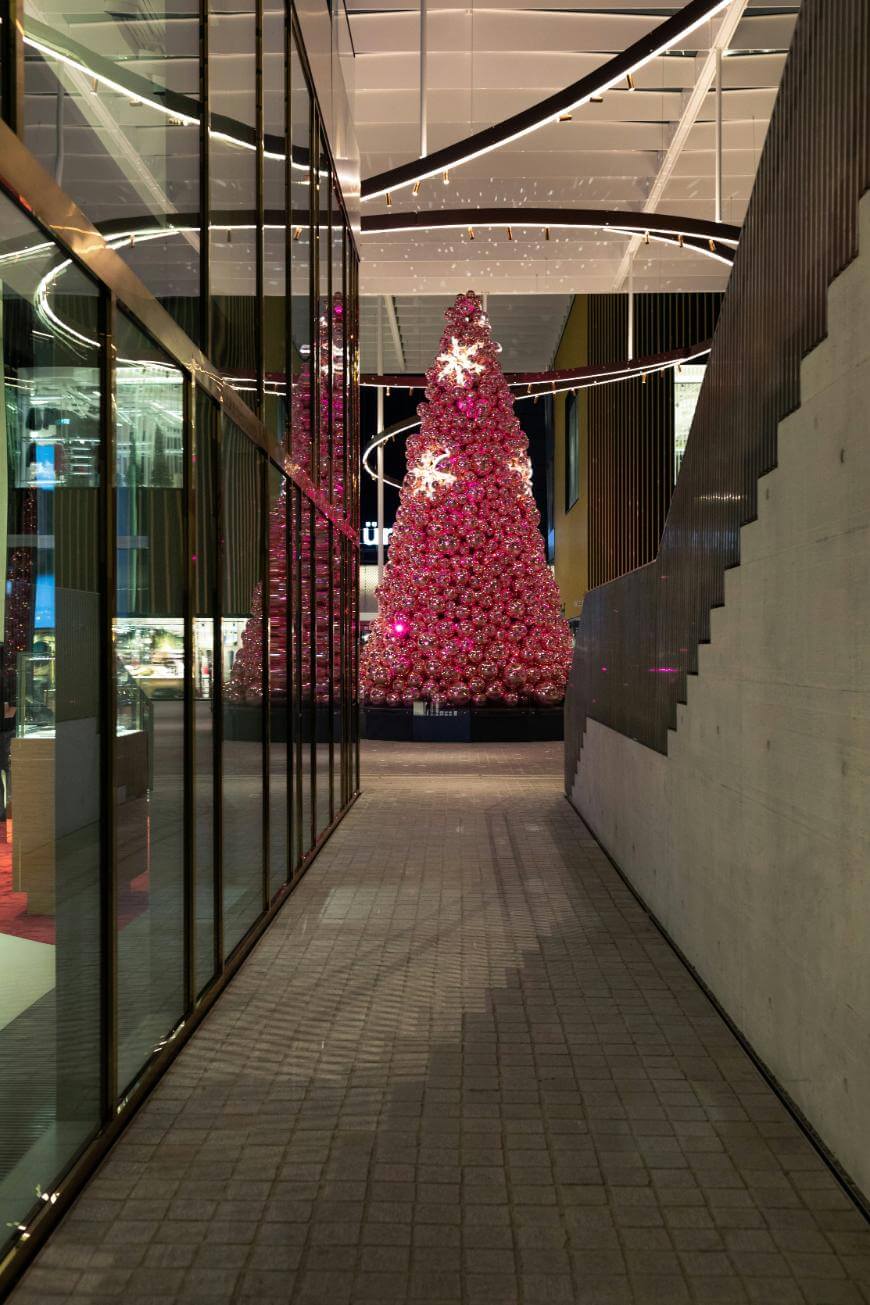 A large pink Christmas tree decorated with lights and stars stands in a modern indoor space, reflected in the glass wall on the left. The area features sleek architecture and a concrete staircase.