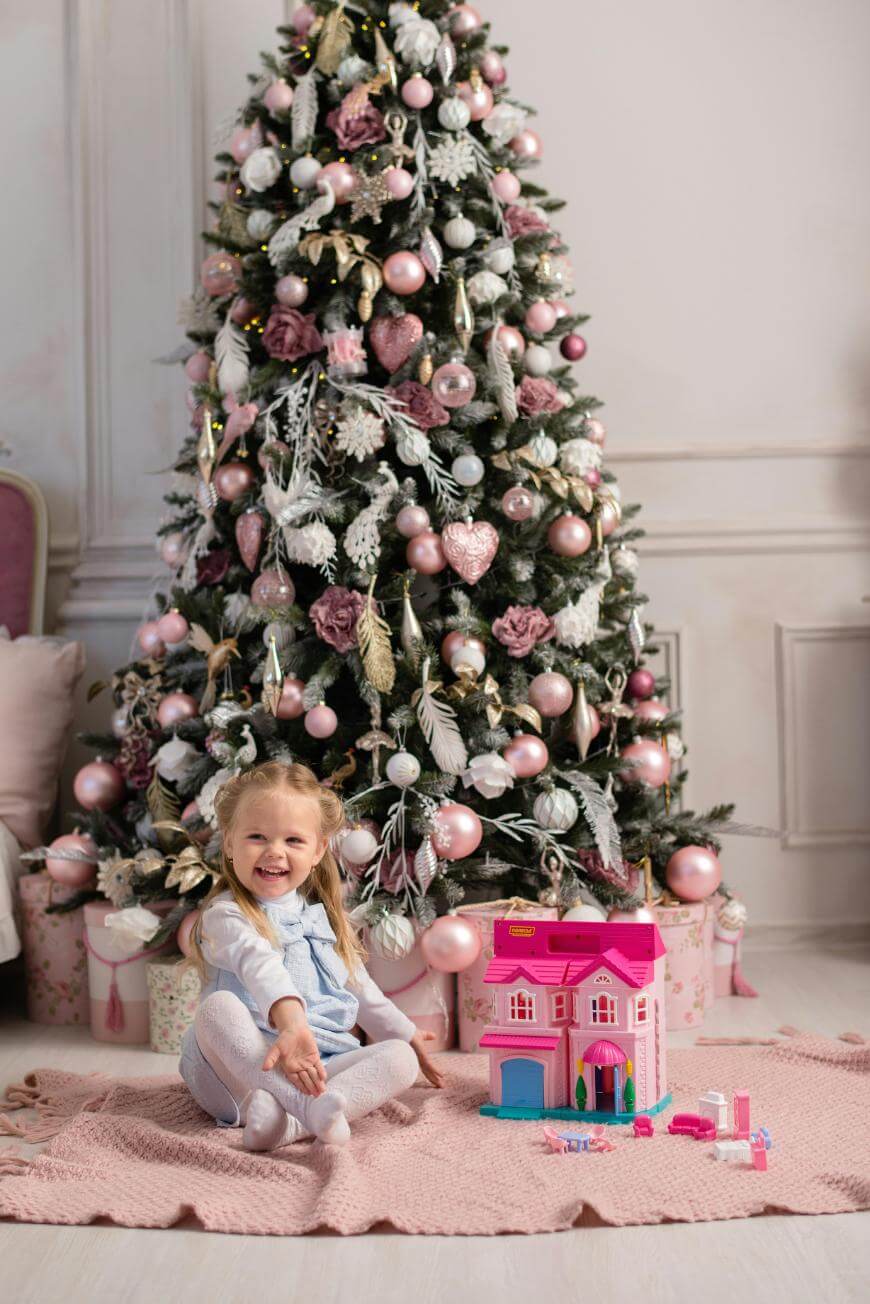 A young girl sits smiling on a pink rug in front of a decorated Christmas tree, surrounded by presents and a pink toy house with accessories. The room has soft pastel colors and festive holiday decor.