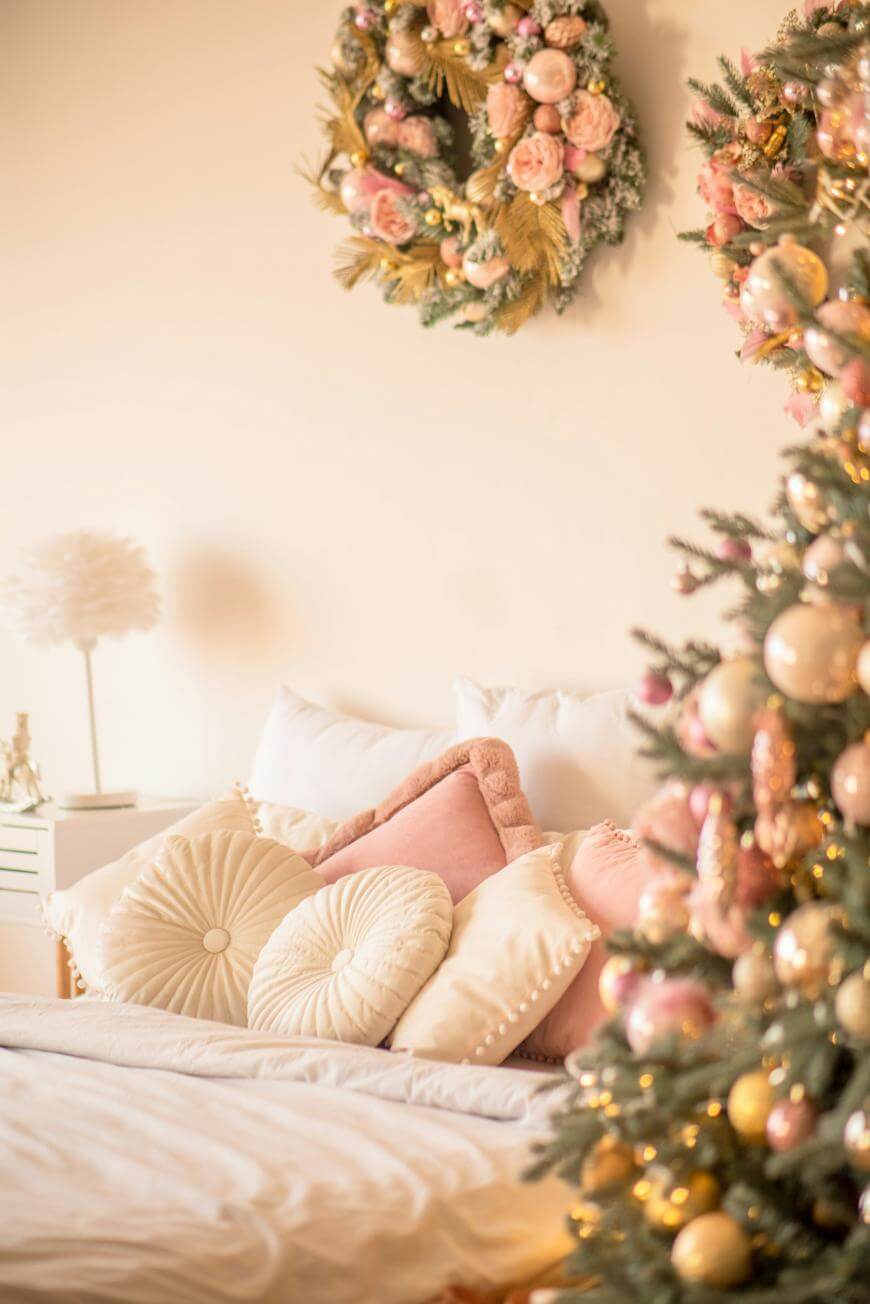 A cozy bedroom decorated for Christmas with pastel pink and gold ornaments on a tree, a floral wreath on the wall, and an arrangement of soft, round pillows on a neatly made bed.