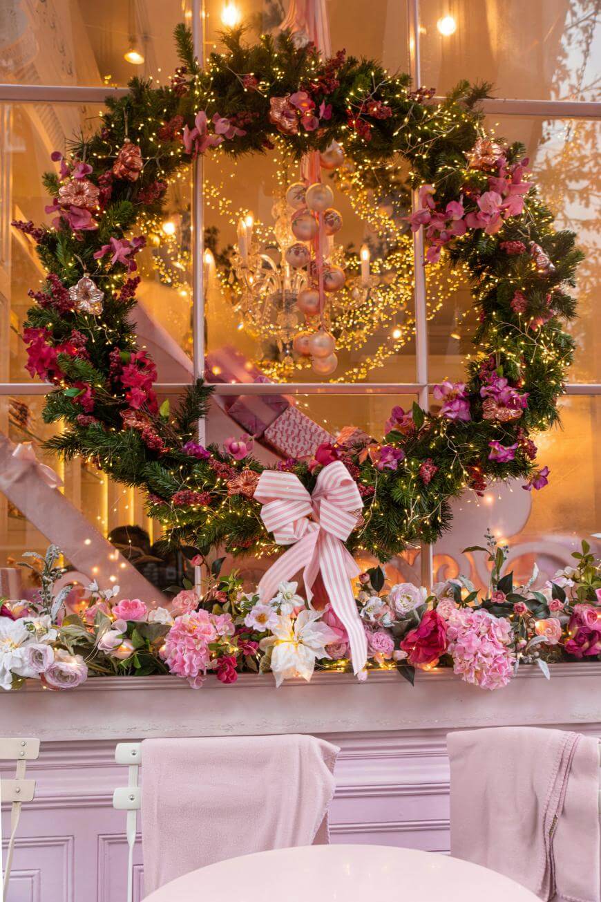 A large, festive wreath with pink and purple flowers, lights, and a striped pink bow decorates a window of a pastel café, with floral garland and soft pink towels draped over chairs outside.
