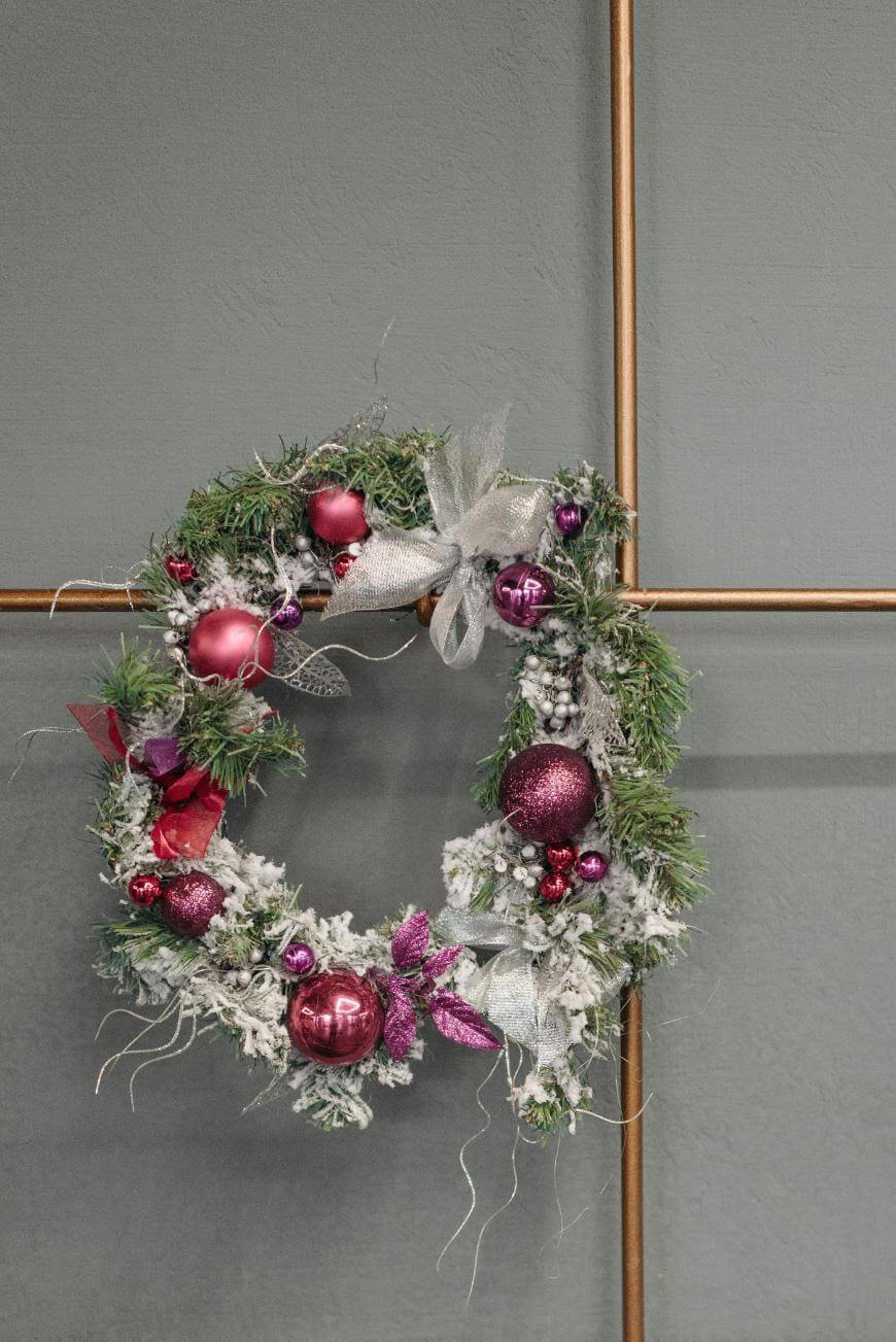 A Christmas wreath decorated with silver ribbon, pink and purple ornaments, and artificial snow hangs on a wall with a metallic grid design in the background.