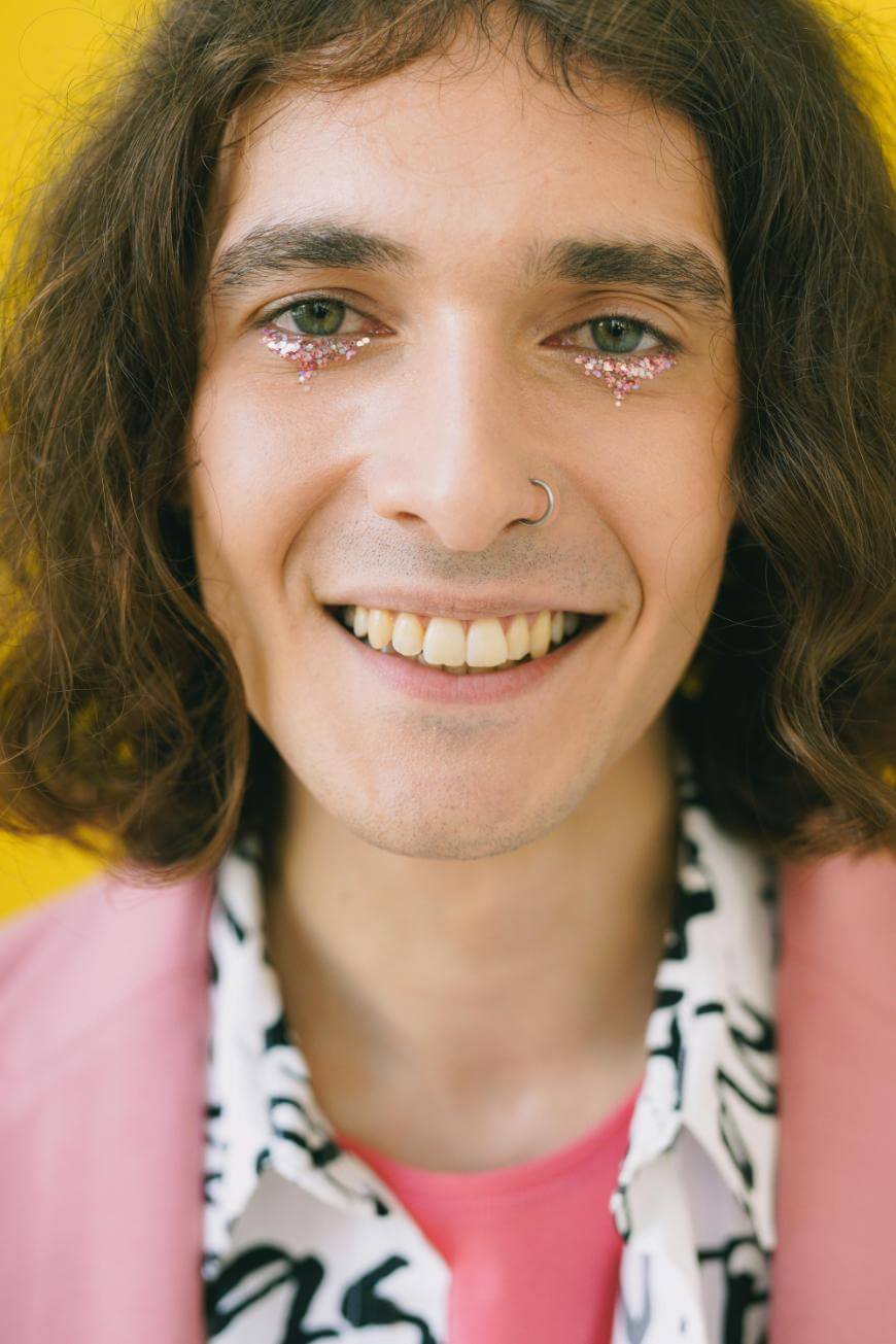 A person with shoulder-length brown hair, a nose ring, and glittery pink makeup under their eyes smiles against a bright yellow background, wearing a pink blazer over a patterned shirt.
