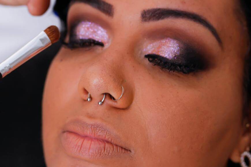 A close-up of a person with closed eyes, wearing shimmery pink eyeshadow and dark eye makeup. A brush is being applied to the eyelid. The person has piercings on their nose and natural, full lips.