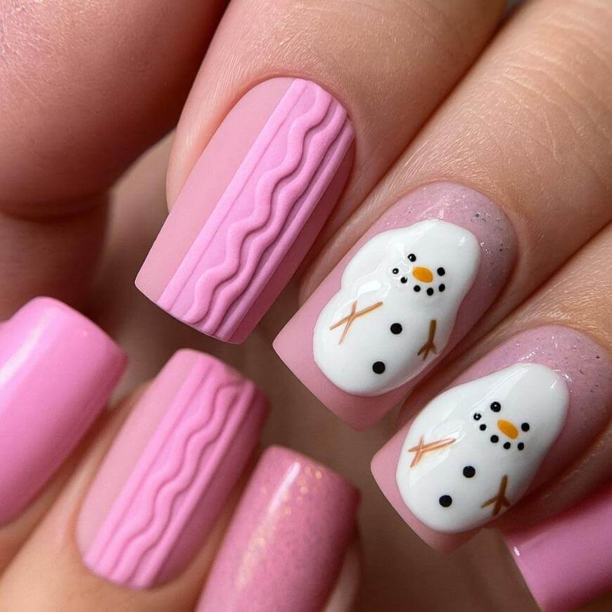 Close-up of a hand with pink winter-themed nail art; two nails have textured wavy lines and two feature snowmen designs with orange noses, black eyes, buttons, and brown stick arms.