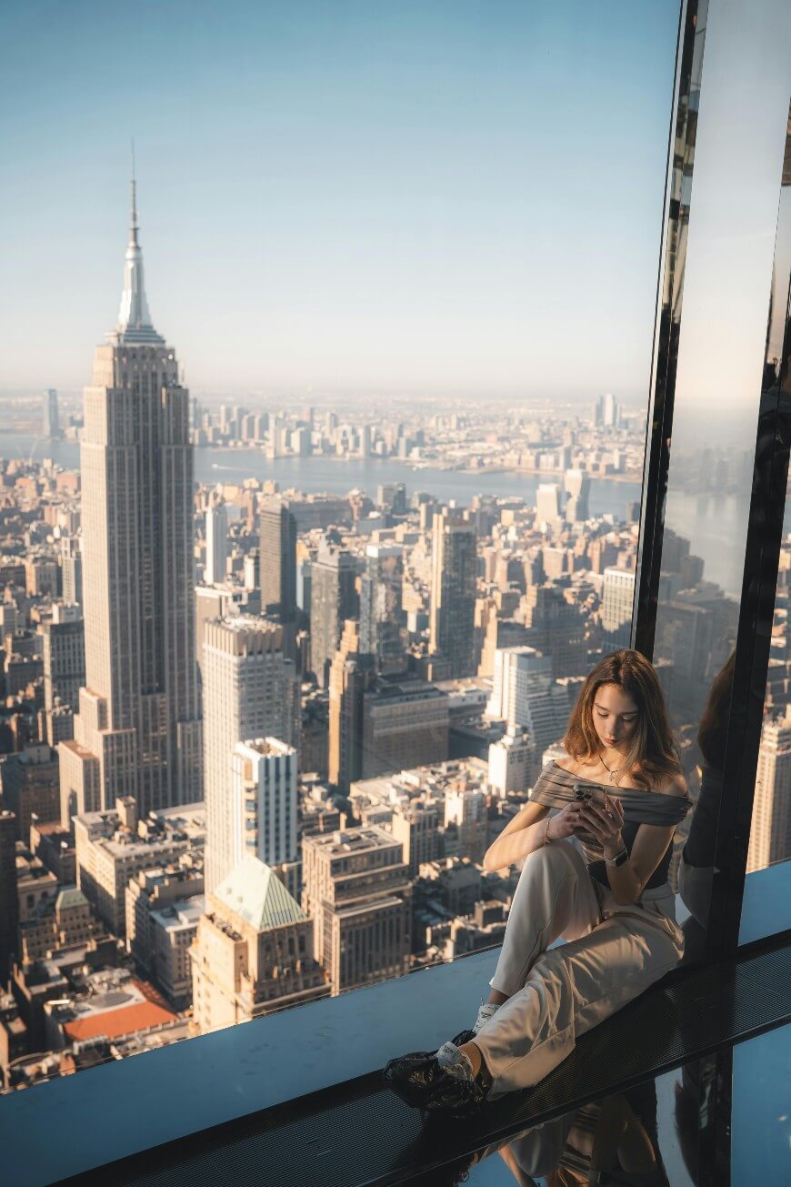A woman sits by a large window overlooking the New York City skyline, with the Empire State Building prominent in the background on a sunny day. She appears relaxed, looking at her phone.