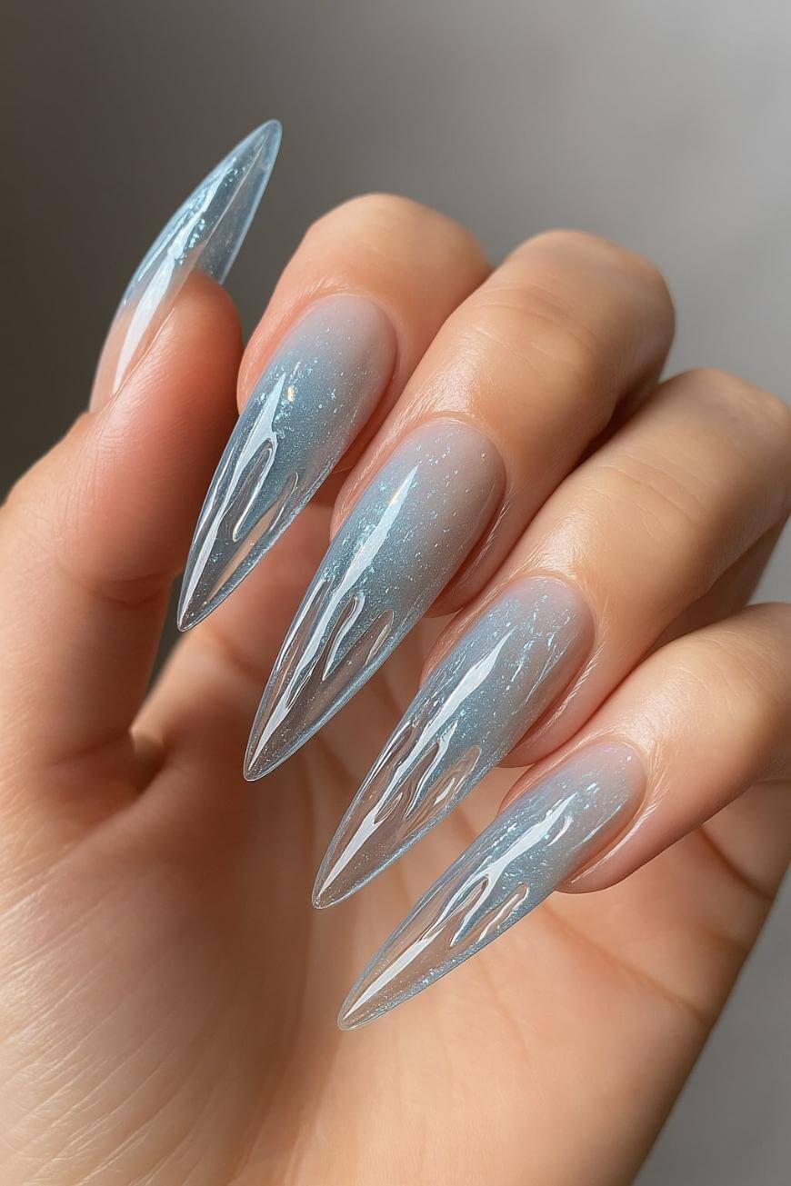 A hand with long, stiletto-shaped nails featuring a glossy, icy blue ombré design with white, flame-like accents and a shimmer finish.