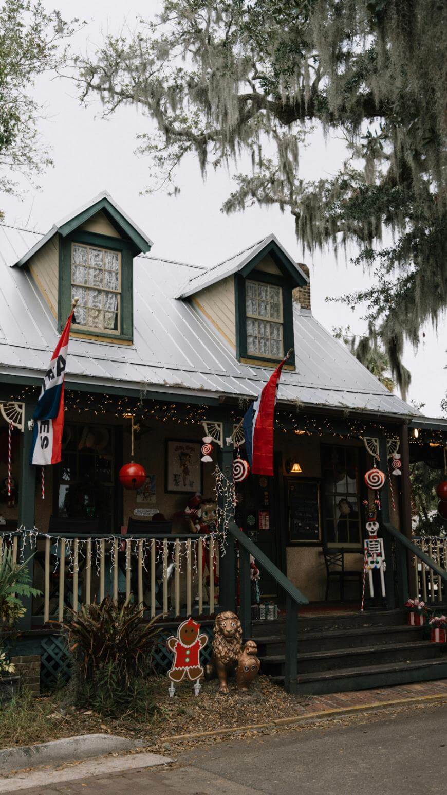 A cozy house decorated for Christmas with string lights, festive flags, candy cane decorations, and a gingerbread man cutout on the porch, surrounded by greenery and hanging Spanish moss.