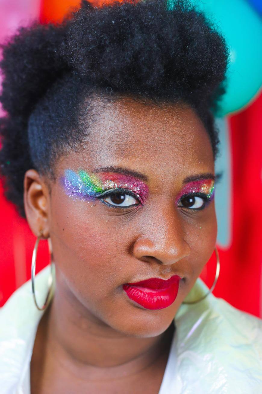 A person with short, curly hair and hoop earrings wears bright red lipstick and bold rainbow glitter eye makeup, set against a colorful, blurred background.