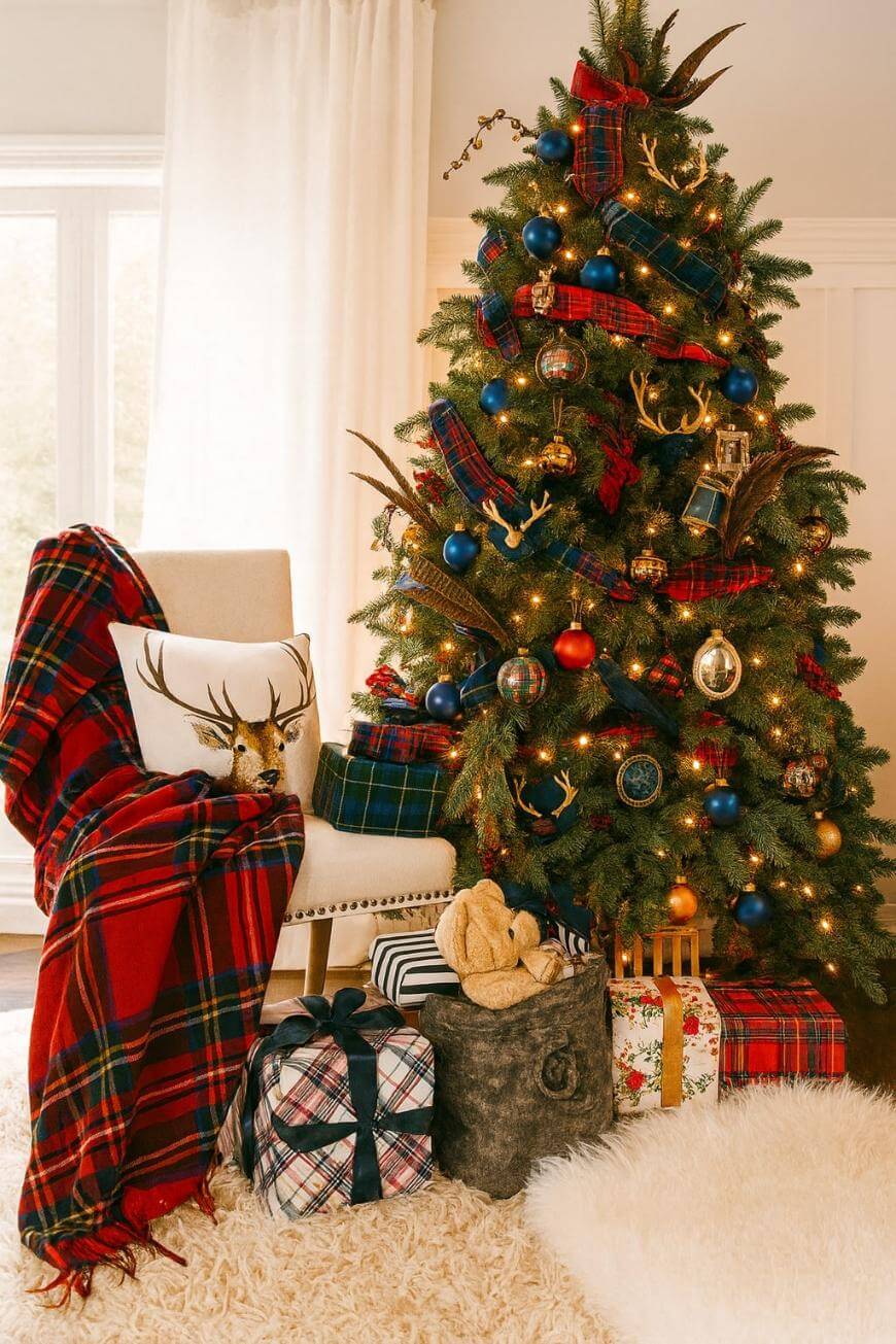 A Christmas tree decorated with blue and red plaid ornaments, deer antlers, and gold lights stands beside gifts wrapped in plaid and neutral paper. A white chair with a stag pillow and plaid blanket sits nearby on a fluffy rug.