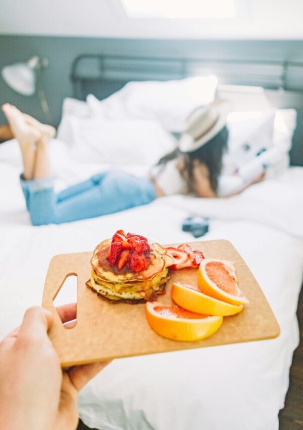 A hand holds a wooden board with pancakes topped with strawberries and orange slices. In the background, a person in jeans and a hat lies on a bed reading a book.