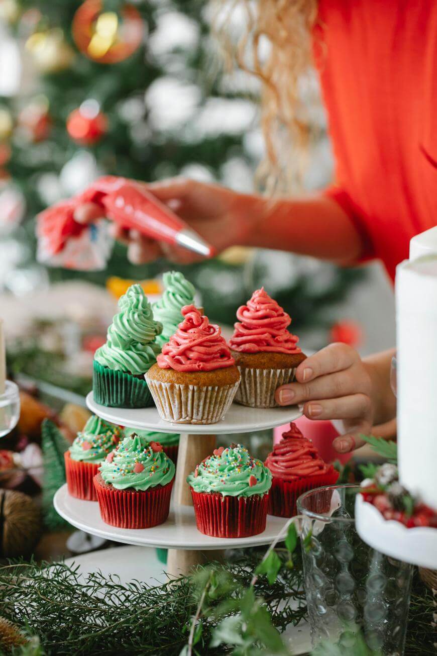 A person decorates festive cupcakes with green and red frosting, placing them on a tiered stand. A Christmas tree is visible in the background, creating a holiday atmosphere.