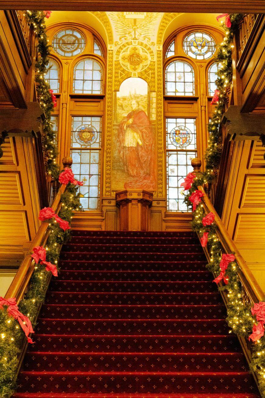 A grand staircase with a red carpet, decorated with garlands and red bows, leads up to stained glass windows and a large mural at the top, all illuminated by warm lighting.