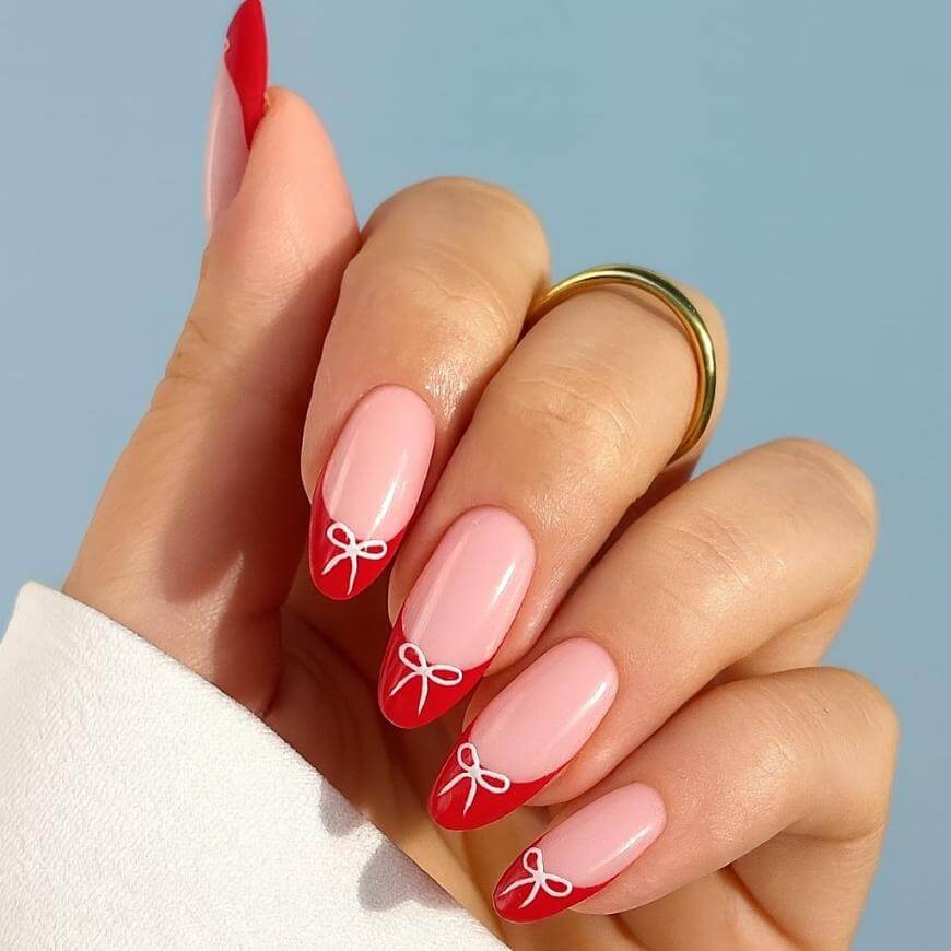 A hand with almond-shaped nails painted light pink, featuring red tips and small white bow designs near the tips. The person is wearing two gold rings and a white sleeve is visible. The background is light blue.