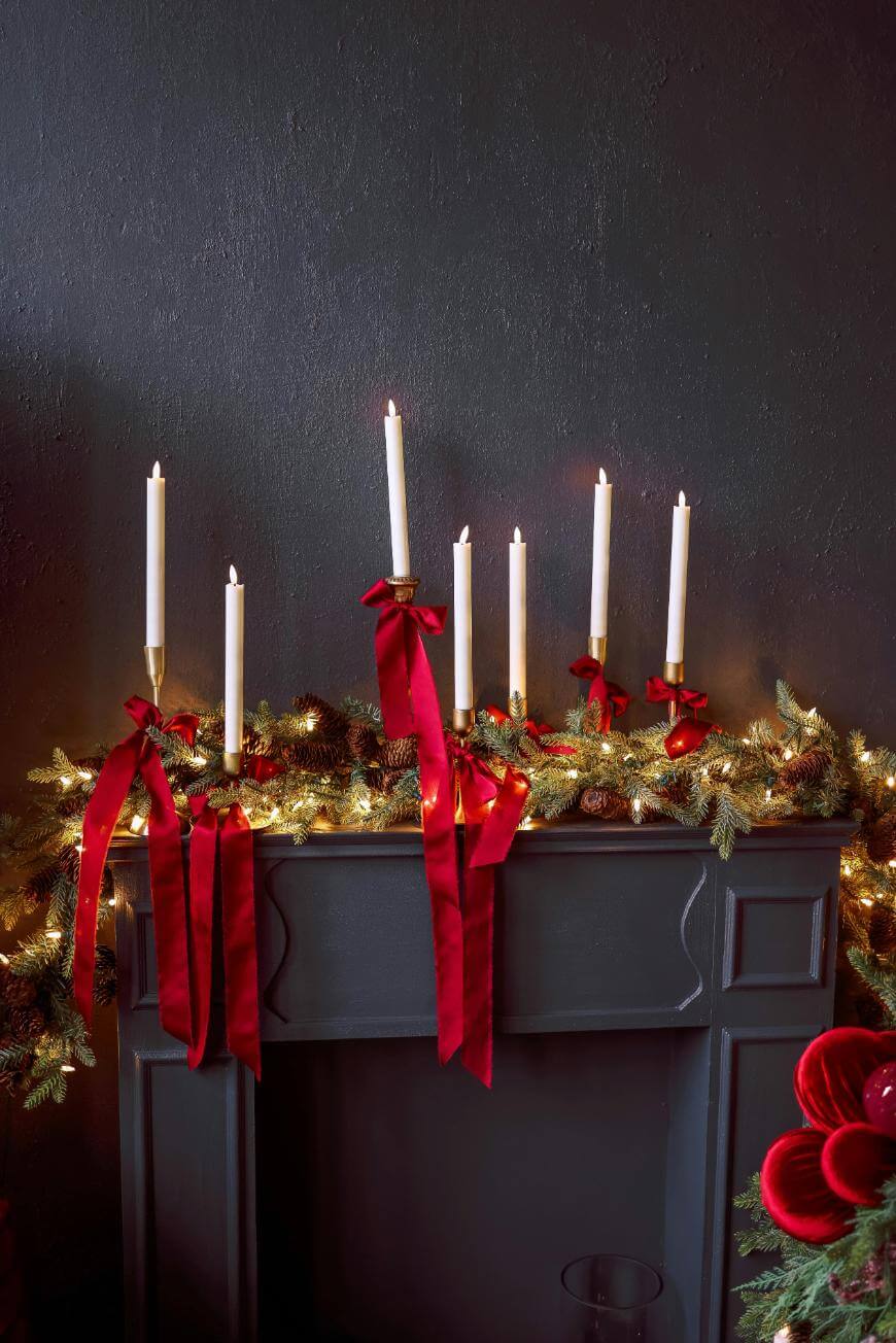 A dark gray mantel decorated with lit white candles, red ribbon bows, pine garland, and string lights, creating a festive holiday display against a dark wall.