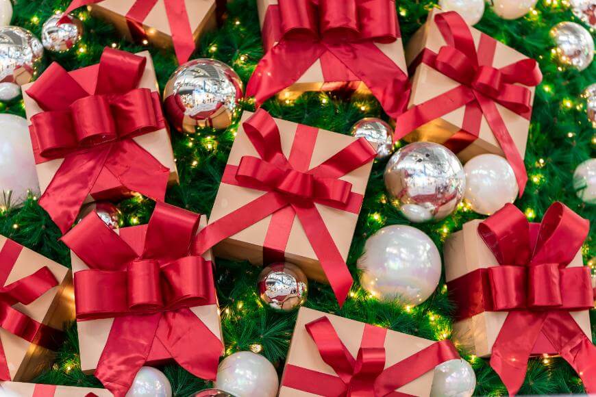Gift boxes wrapped in brown paper with red ribbons are arranged on green pine branches, surrounded by shiny silver and white ornaments and twinkling lights, creating a festive holiday display.