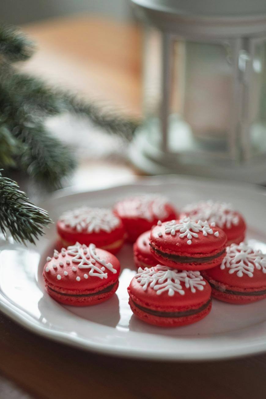 A white plate with red macarons decorated with white snowflake icing, placed near pine branches and a blurred white lantern in the background.