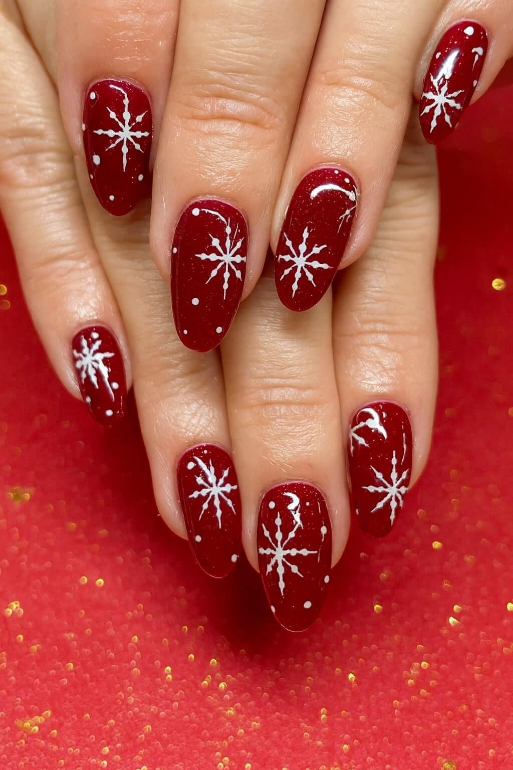 A close-up of hands with red nail polish, each nail decorated with white snowflake designs and small white dots, creating a festive winter look. The background is red with gold sparkles.