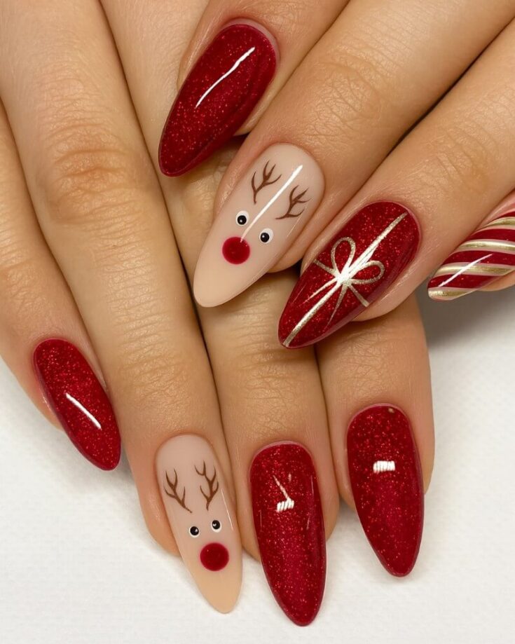 A hand with festive nail art: most nails are painted sparkly red, one has a reindeer face with a red nose on a nude base, and another has white ribbon and candy cane designs on red.