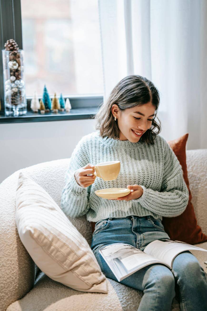 A young woman in a light blue sweater sits on a cozy chair, smiling as she holds a yellow teacup and saucer, with an open book on her lap. A window with winter-themed decorations is in the background.