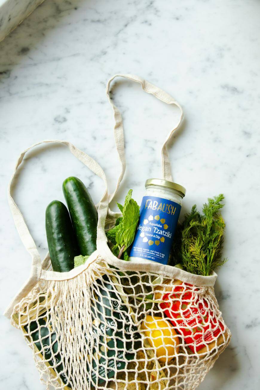 A reusable mesh grocery bag with cucumbers, leafy greens, a yellow bell pepper, a red bell pepper, and a jar of vegan tzatziki sits on a marble countertop.
