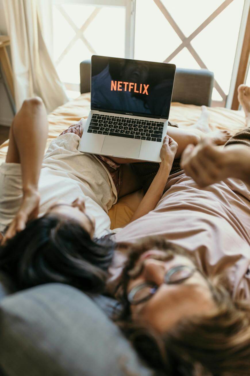A couple lies on a bed, relaxing together while watching Netflix on a laptop. The screen displays the Netflix logo, and the setting appears cozy and casual with natural light coming in.