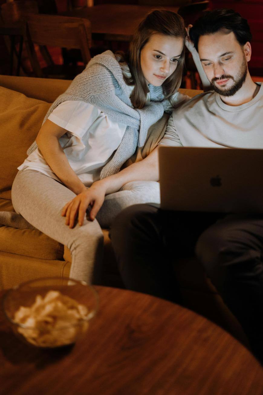 A woman and a man sit closely together on a couch, looking at a laptop screen. The woman has her arm around the man’s shoulder, and a bowl of chips is on a wooden table in the foreground.
