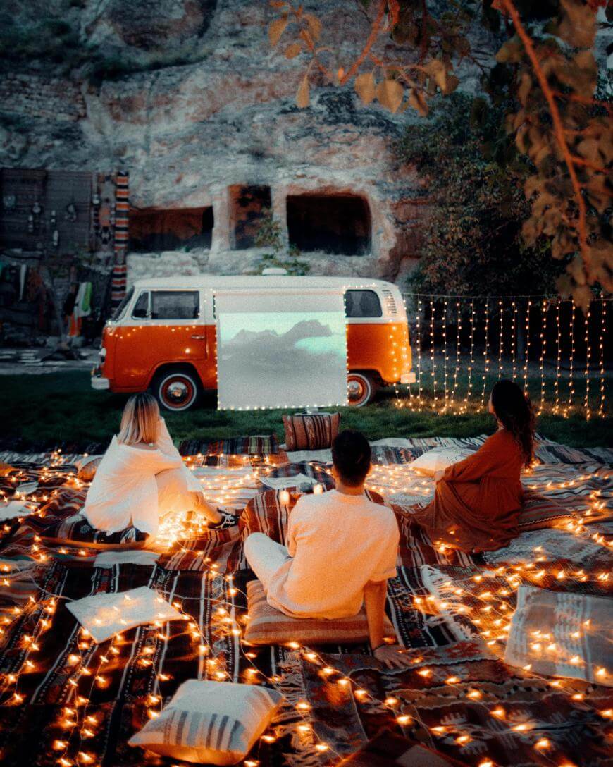 Three people sit on blankets and pillows outdoors at night, watching a movie projected onto a red and white van. String lights and cozy decor create a warm, inviting atmosphere surrounded by nature.