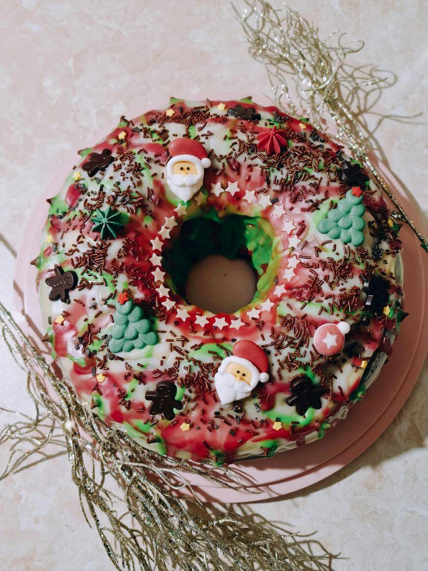 A festive Christmas wreath cake decorated with white icing, chocolate sprinkles, and holiday-themed candies shaped like Santa, trees, stars, and gingerbread men, set on a pink plate with gold tinsel.