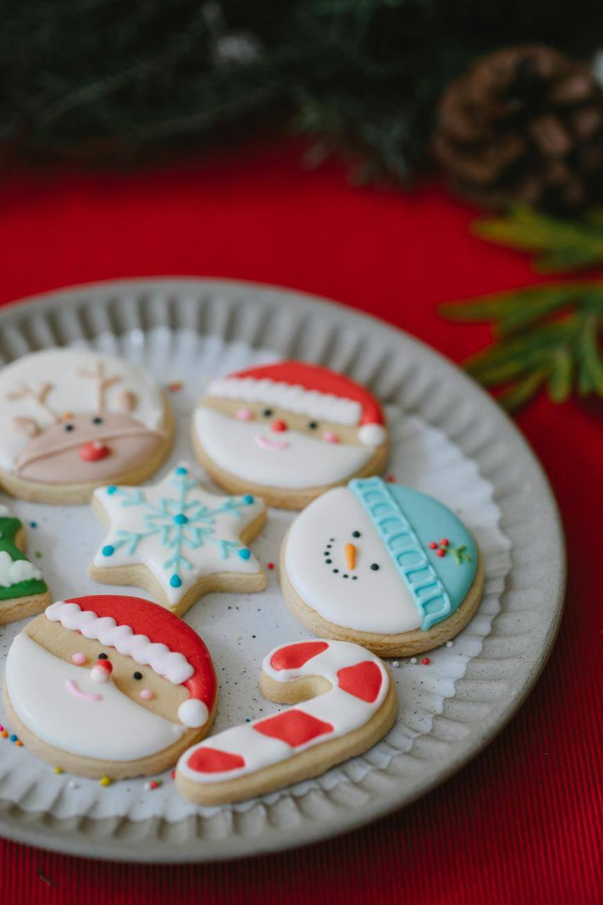 A plate with decorated Christmas cookies, including a reindeer, Santa faces, a snowman, a snowflake, and a candy cane, on a red surface with pine and greenery in the background.