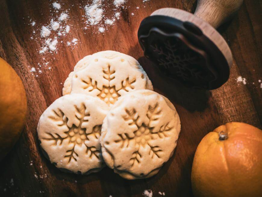 Three round cookies with snowflake patterns are on a wooden surface, surrounded by scattered flour, two oranges, and a wooden cookie stamp.