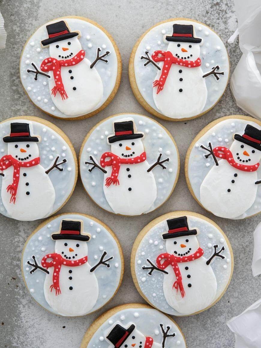 Round cookies decorated with snowmen made of white icing, red scarves, black hats, orange carrot noses, and stick arms. The background icing is light blue with white dot details resembling snow.