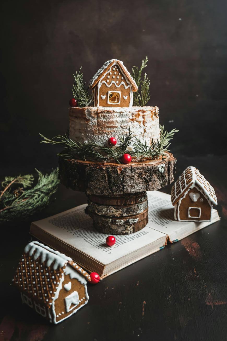 A festive cake topped with a small gingerbread house, greenery, and red berries sits on a rustic wooden stand placed on an open book, with two more gingerbread houses beside it.
