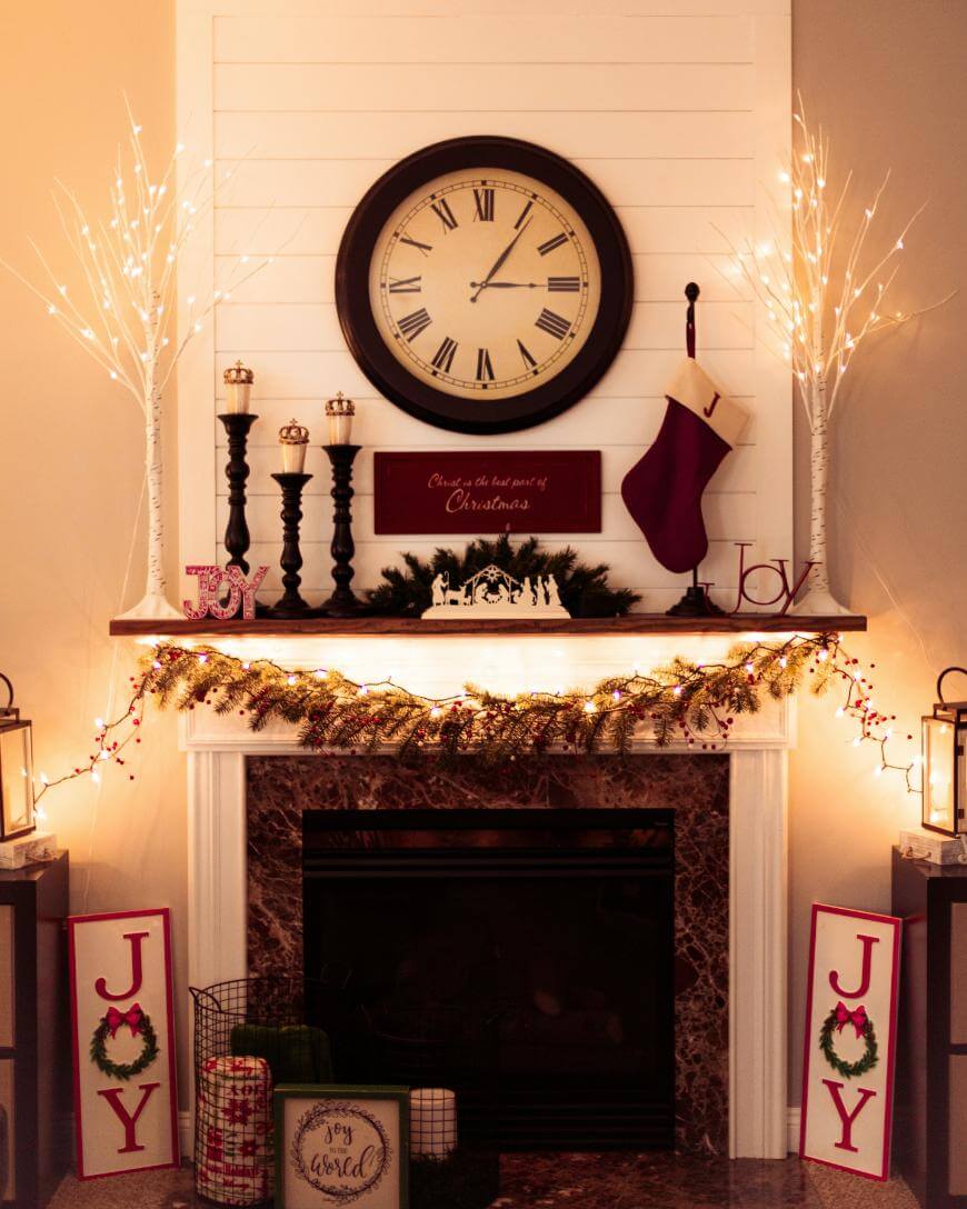 A decorated fireplace with garland and string lights, a large clock above the mantel, two JOY signs on the floor, candles, a stocking, and Christmas ornaments creating a festive holiday scene.